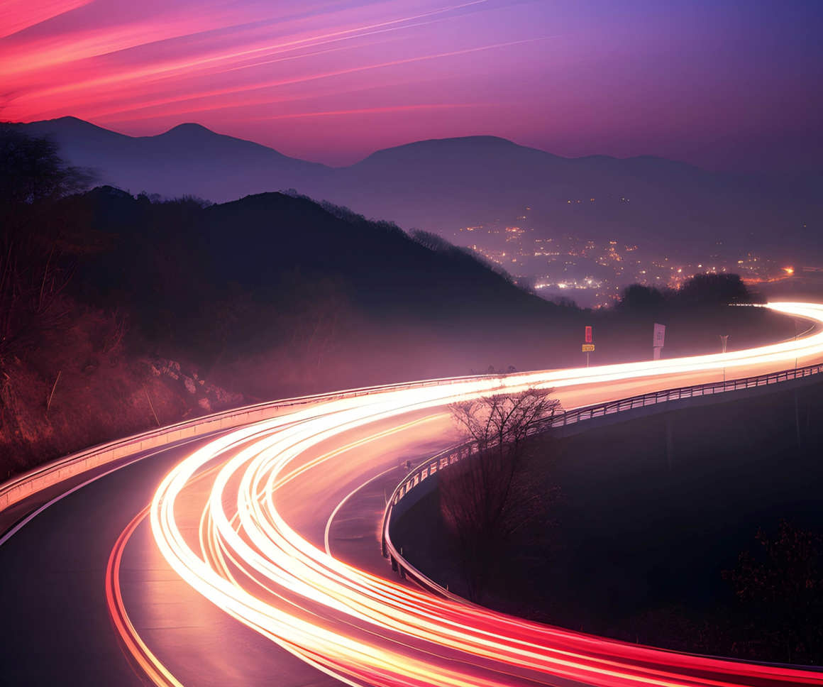 A winding road with bright red and white light trails from vehicles creates a dynamic curve, set against a twilight sky with deep pink and purple hues. Mountains are visible in the distance, and city lights illuminate the horizon.