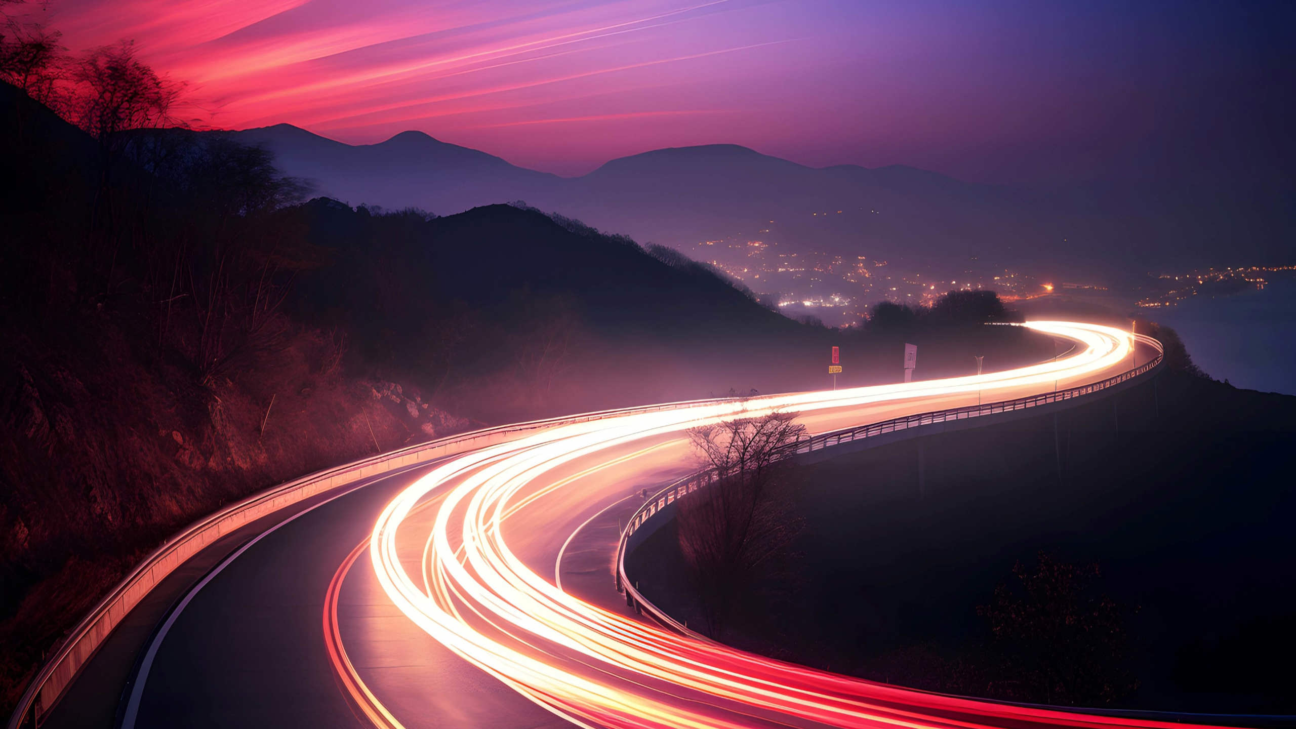 A winding road with bright red and white light trails from vehicles creates a dynamic curve, set against a twilight sky with deep pink and purple hues. Mountains are visible in the distance, and city lights illuminate the horizon.