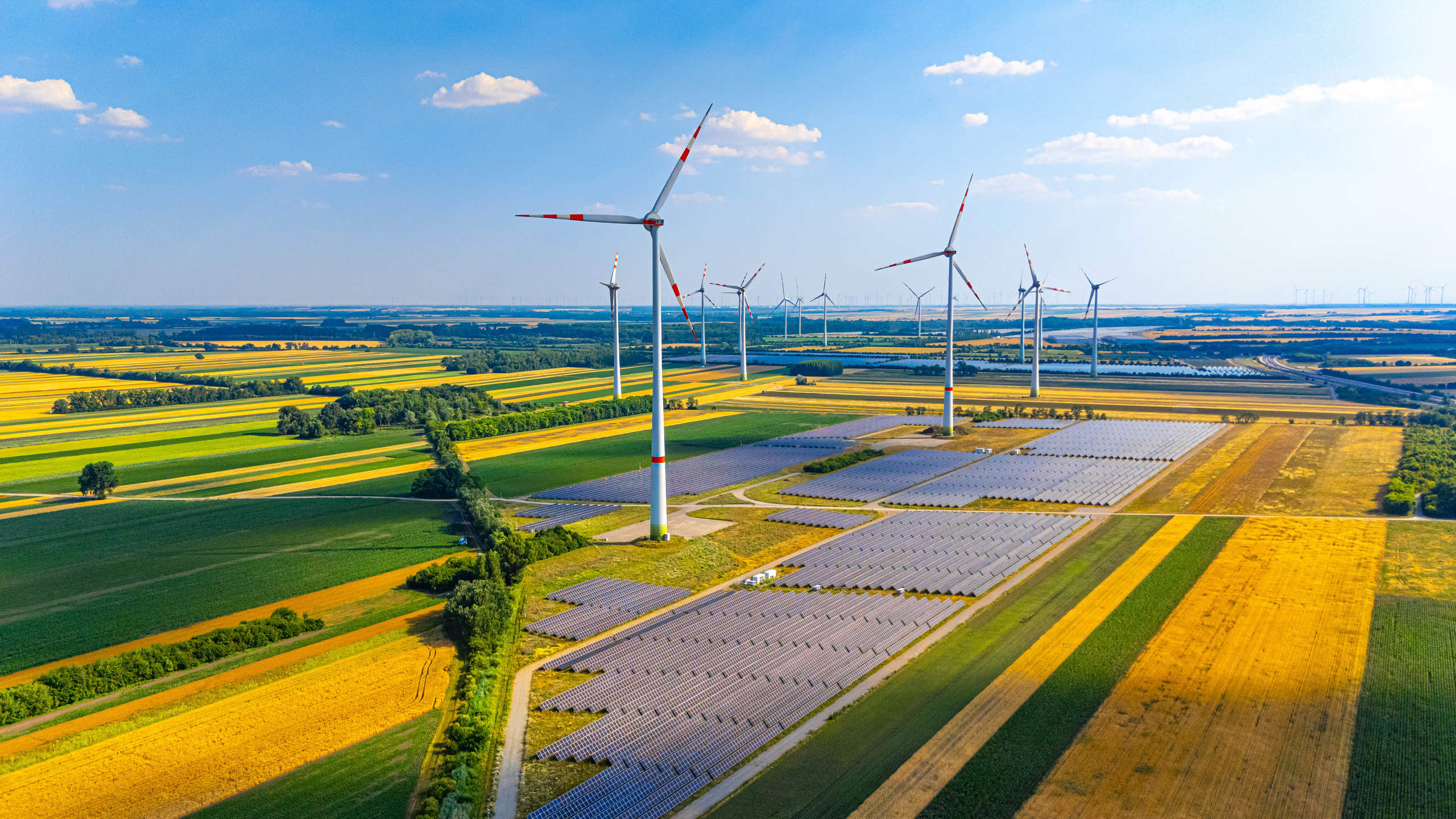 Wind turbines and solar panels in a green field on a sunny day. A large area with numerous wind turbines and solar panels under a clear sky. Fields stretch in different colours.