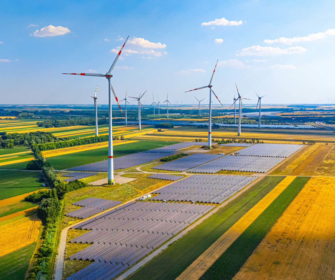 Wind turbines and solar panels in a green field on a sunny day. A large area with numerous wind turbines and solar panels under a clear sky. Fields stretch in different colours.