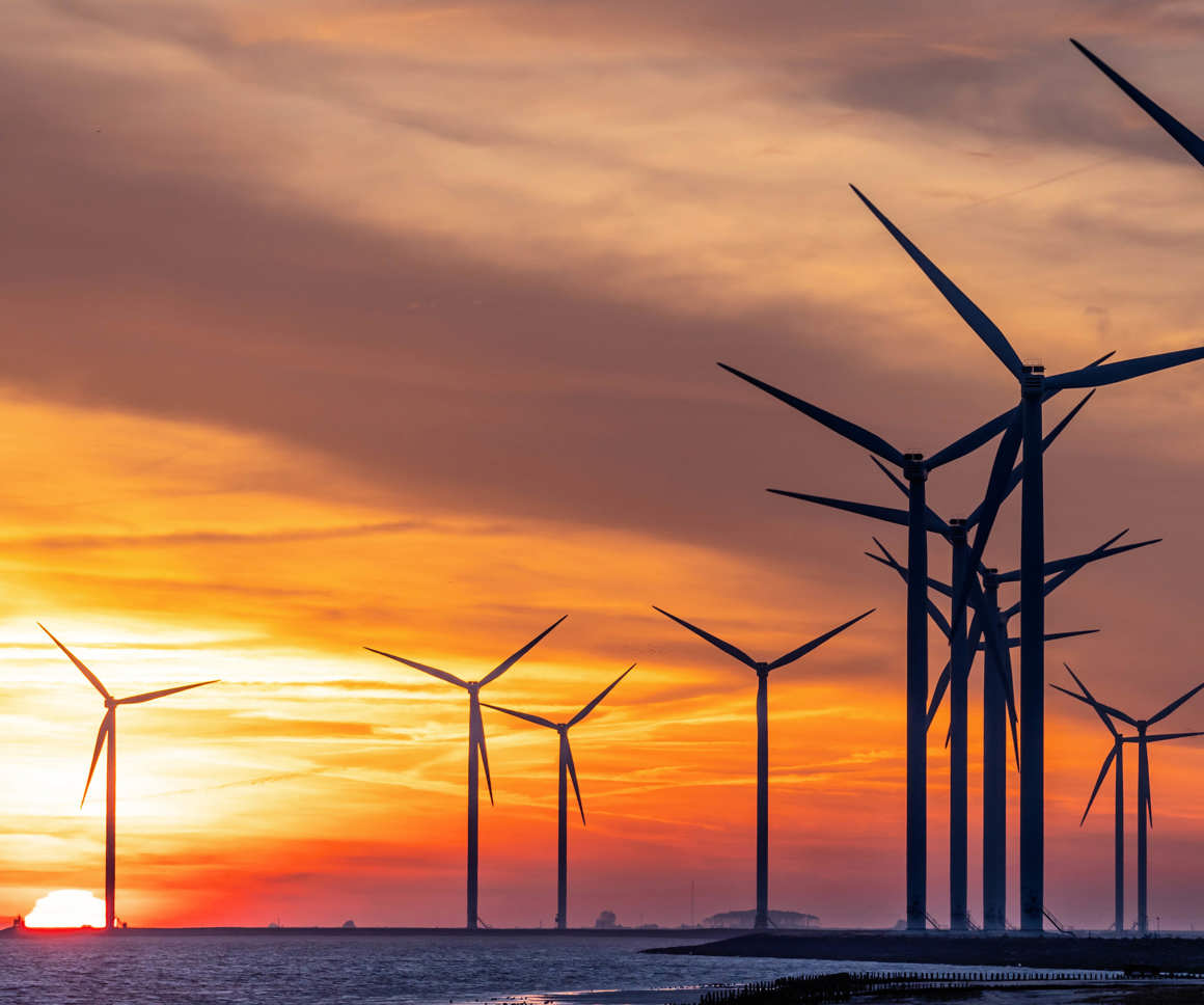 A series of wind turbines silhouetted against a vibrant sunset sky over a body of water. The sun is partially visible near the horizon, casting an orange and yellow glow across the scene.