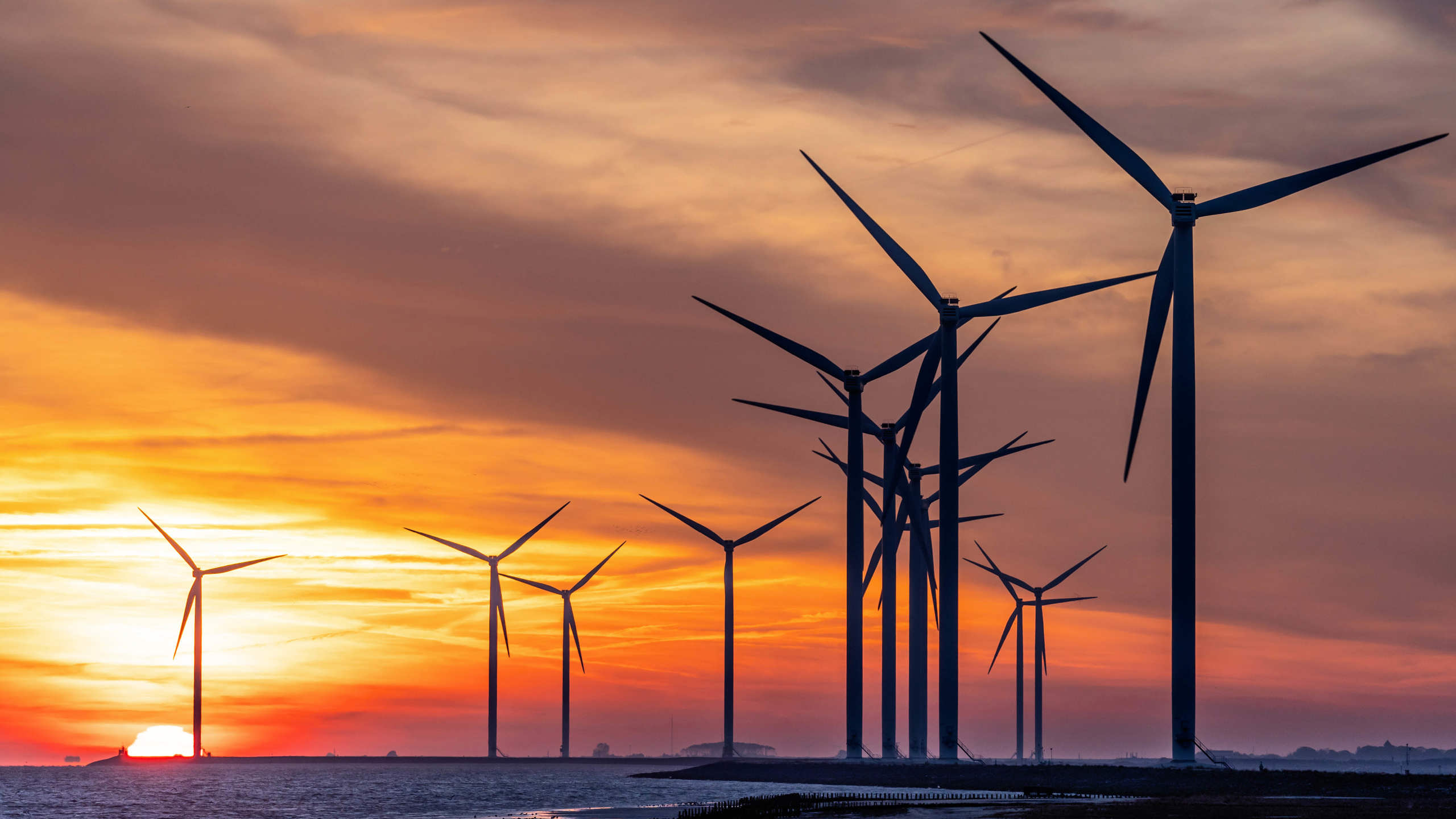 A series of wind turbines silhouetted against a vibrant sunset sky over a body of water. The sun is partially visible near the horizon, casting an orange and yellow glow across the scene.